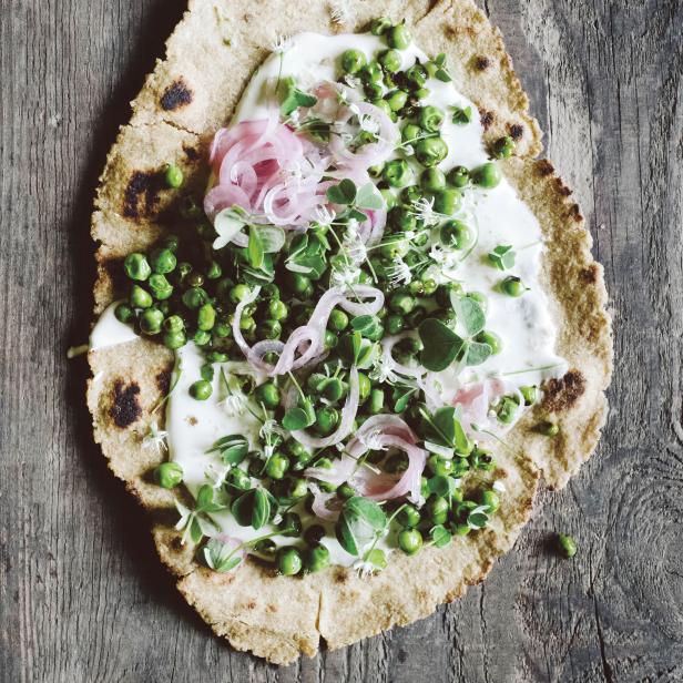 Ein Fladenbrot belegt mit Erbsen, eingelegten Zwiebeln und Kräutern auf einem Holztisch.