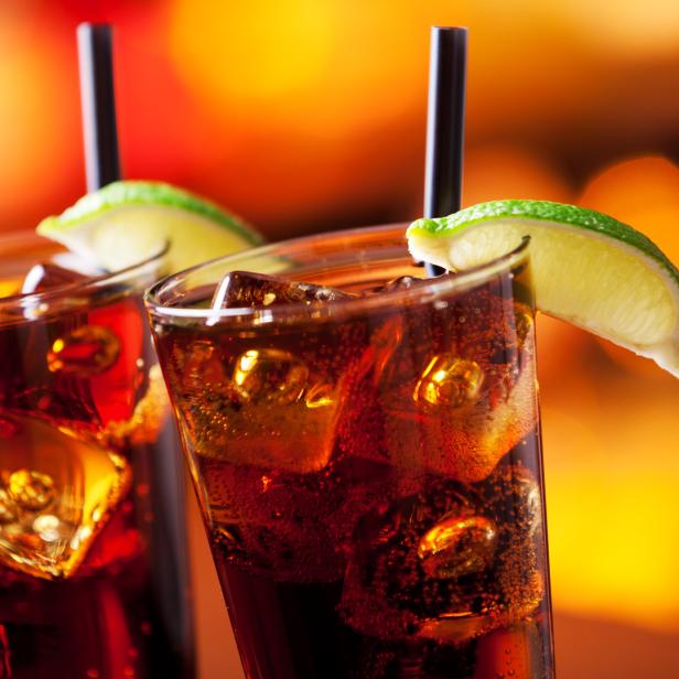 Zwei Gläser mit Cola, Eis und Limetten vor einem verschwommenen, orangefarbenen Hintergrund.