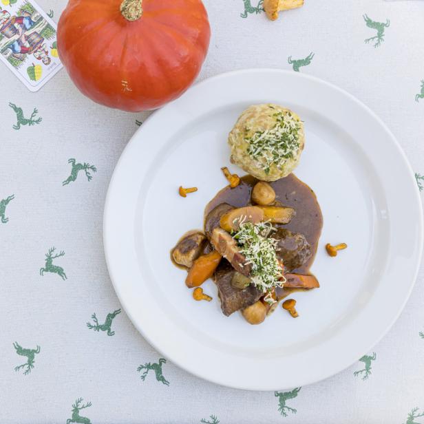 Ein Teller mit Wildragout, Semmelknödel und Pfifferlingen auf einer Tischdecke mit Hirschmuster.