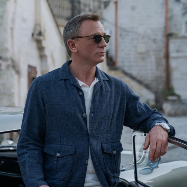 Daniel Craig als James Bond posiert an einem Oldtimer vor einem Gebäude.