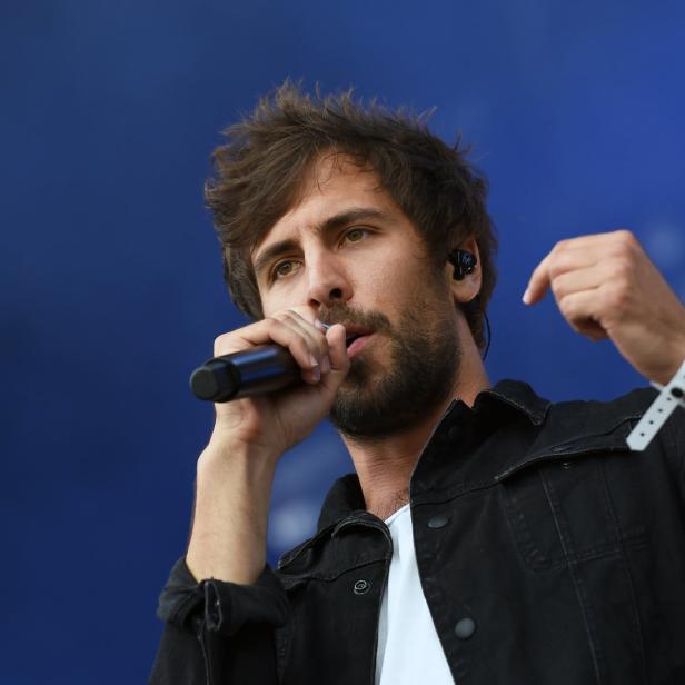 Ein Mann mit Bart singt in ein Mikrofon vor einem blauen Hintergrund.