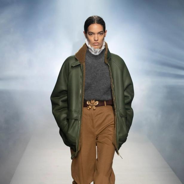 Eine Frau auf dem Laufsteg trägt eine grüne Jacke, braune Hose und Stiefel mit Schlangenmuster.