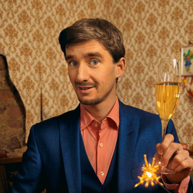 Ein Mann im Anzug hält ein Glas Sekt und ein Wunderkerze in die Kamera.