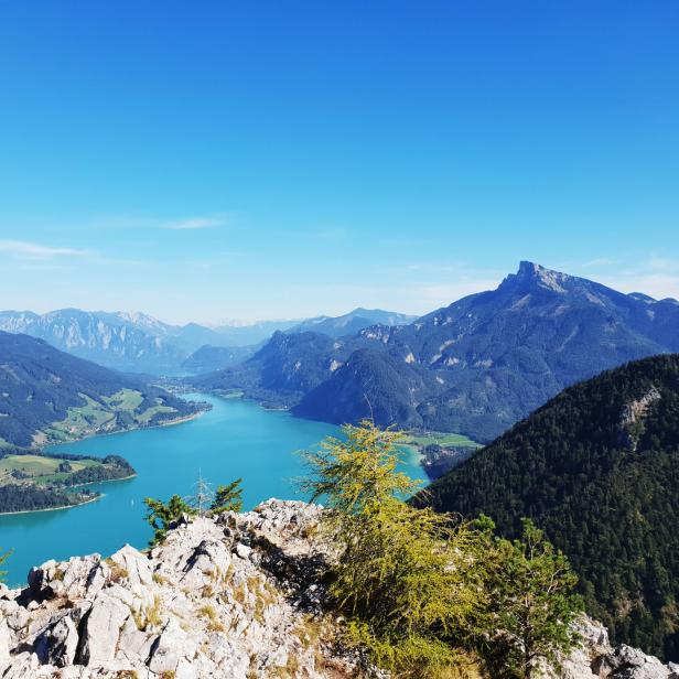 Blick von einem felsigen Gipfel auf einen türkisfarbenen See und bewaldete Berge unter blauem Himmel.