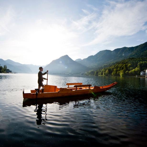 Ein Mann in Tracht stakt ein traditionelles Holzboot auf einem Bergsee.