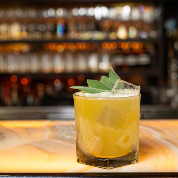 Ein gelber Cocktail mit Eis und Salbeiblättern auf einer Bar.