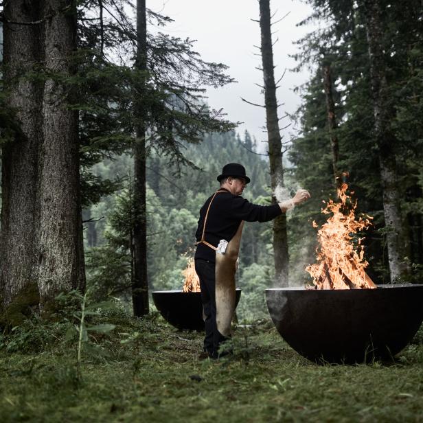 Ein Mann mit Hut steht im Wald und hantiert an einem Feuer in einer Feuerschale.