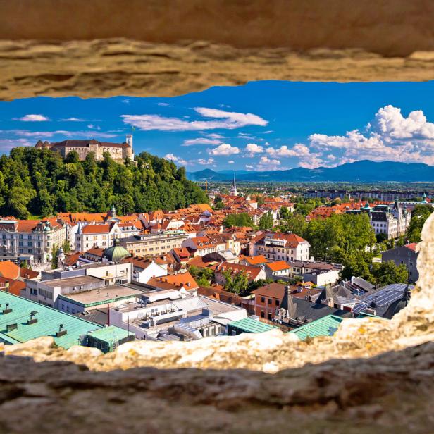 Blick durch eine Maueröffnung auf die Stadt Ljubljana mit Burg im Hintergrund.
