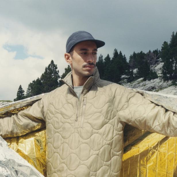 Ein Mann mit Mütze steht in einer Berglandschaft, gehüllt in eine goldene Rettungsdecke.
