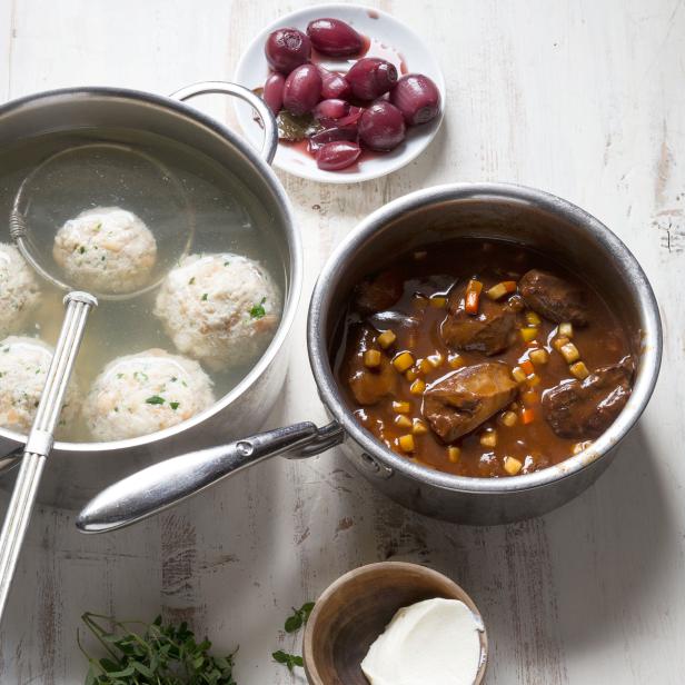 Ein Topf mit Knödeln, ein Topf mit Gulasch, eingelegte Zwiebeln und frischer Majoran auf einem Tisch.