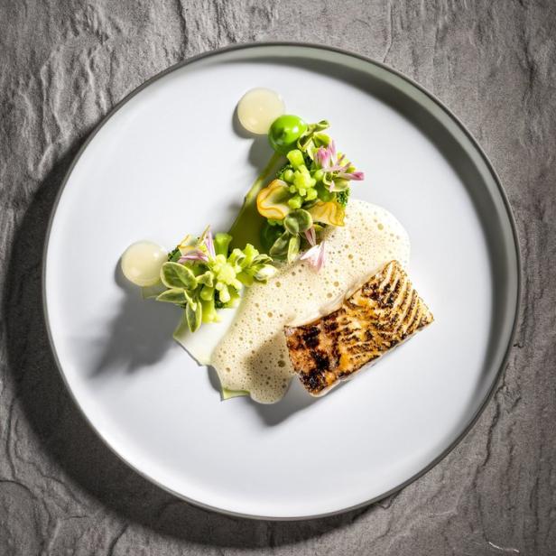 Ein Teller mit gegrilltem Fisch, Gemüse und einer hellen Soße auf einem grauen Steintisch.