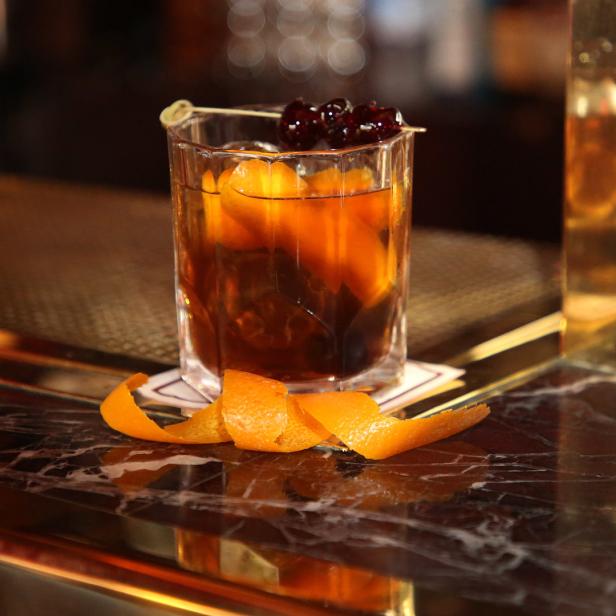 Ein Old Fashioned Cocktail mit Orangenschalen und Kirschen auf einer Marmoroberfläche.
