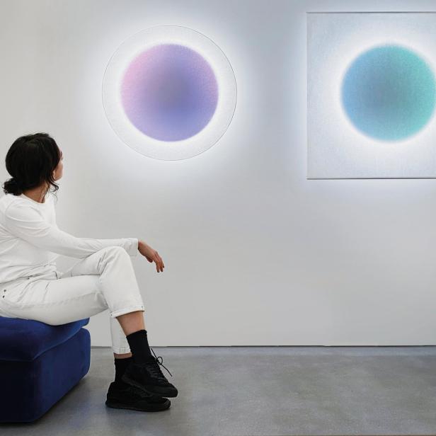 Eine Frau sitzt vor zwei abstrakten, kreisförmigen Kunstwerken in einer Galerie.