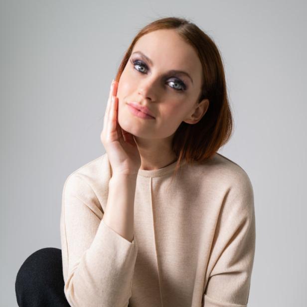 Eine Frau mit roten Haaren posiert in einem beigen Pullover und schwarzen Hosen.