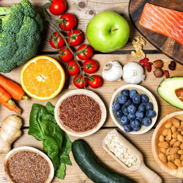 Verschiedene gesunde Lebensmittel wie Gemüse, Obst, Nüsse, Lachs, Reis, Haferflocken und Samen liegen auf einem Holztisch.