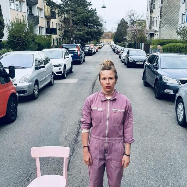 Eine Frau in einem lila Overall steht mit einem rosa Stuhl auf einer Straße.
