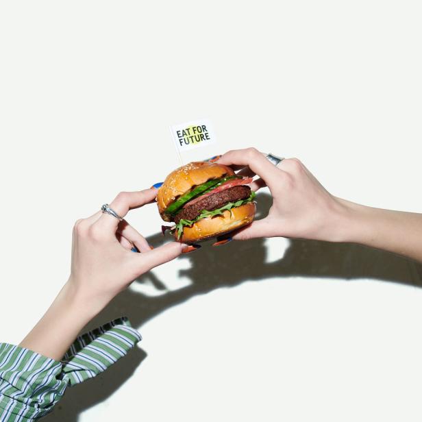 Zwei Hände halten einen Burger mit einem kleinen Fähnchen, auf dem „Eat for Future“ steht.