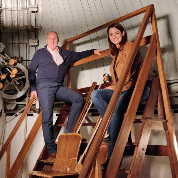 Zwei Personen posieren auf einer Holztreppe vor einem großen Teleskop in einer Sternwarte.