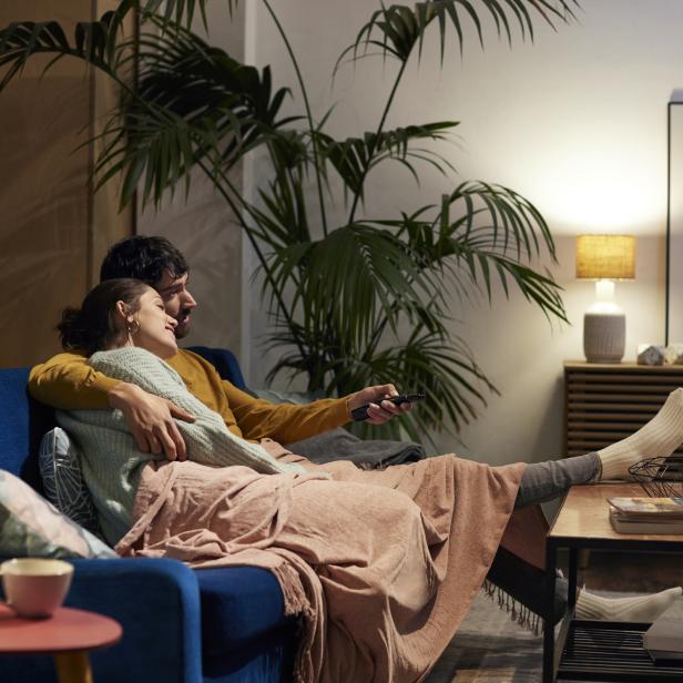 Ein Paar kuschelt sich auf einem blauen Sofa unter einer Decke und schaut fern.