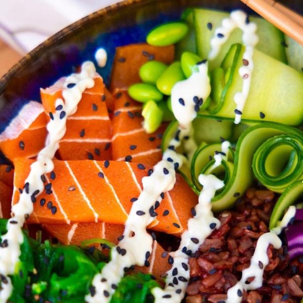 Eine Poke Bowl mit Lachs, Reis, Gurke, Edamame und Algen, garniert mit einer hellen Soße und Sesamsamen.