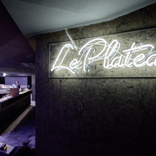 Ein Leuchtschild mit der Aufschrift „Le Plateau“ an einer dunklen Wand in einer Bar.