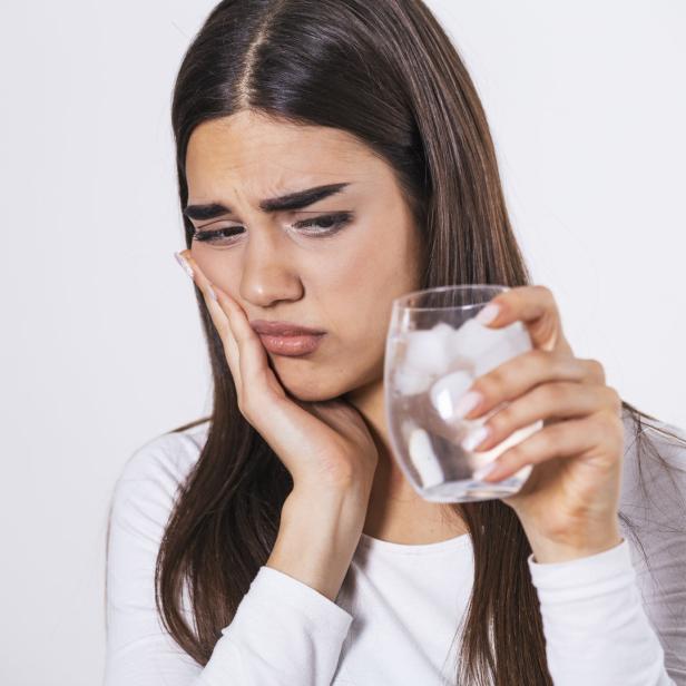 Eine Frau mit Zahnschmerzen hält ein Glas Eiswasser an ihre Wange.