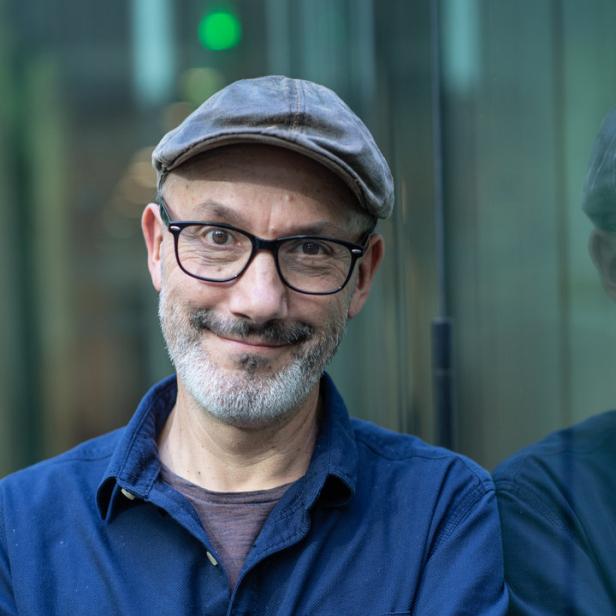 Ein Mann mit Brille und Schirmmütze steht mit verschränkten Armen vor einer Glaswand.