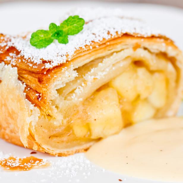 Ein Stück Apfelstrudel mit Puderzucker und Vanillesauce auf einem Teller.