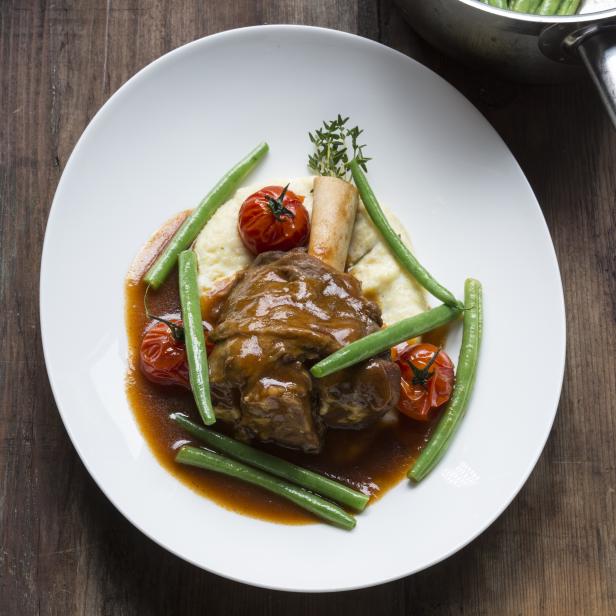 Ein Teller mit Lammhaxe, Kartoffelpüree, grünen Bohnen und Tomaten auf einem Holztisch.