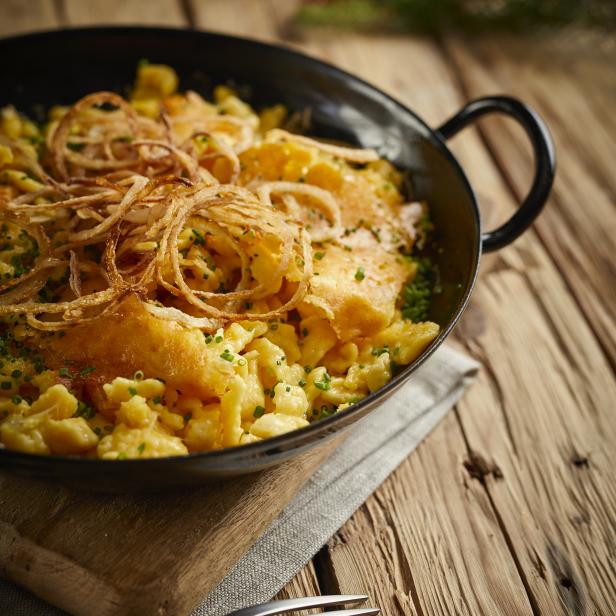 Käsespätzle mit Röstzwiebeln in einer schwarzen Pfanne auf einem Holztisch.