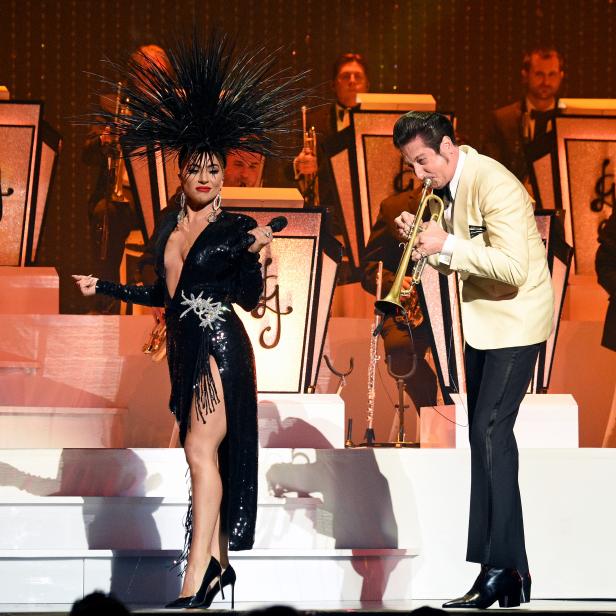 Eine Sängerin mit extravagantem Kopfschmuck und ein Trompeter treten mit einer Band auf einer Bühne auf.