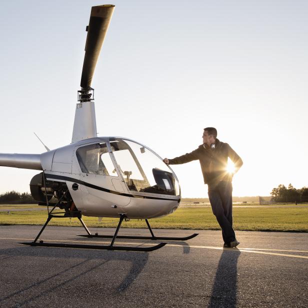 Ein Mann steht neben einem silbernen Hubschrauber auf einem Flugfeld.