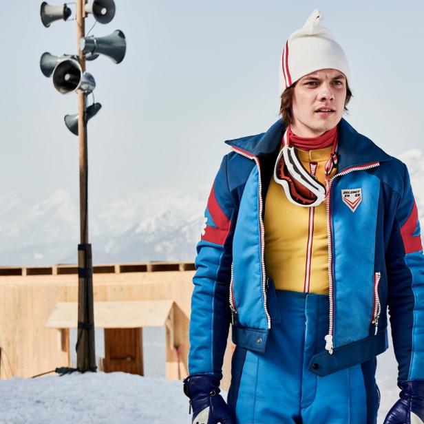 Ein Mann in Skikleidung steht vor einer verschneiten Bergkulisse.