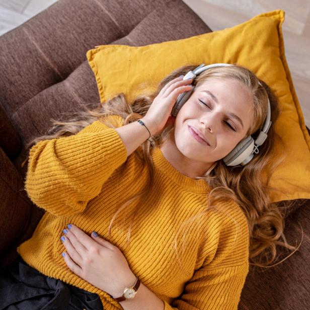 Eine junge Frau liegt entspannt mit Kopfhörern auf einem Sofa und hört Musik.