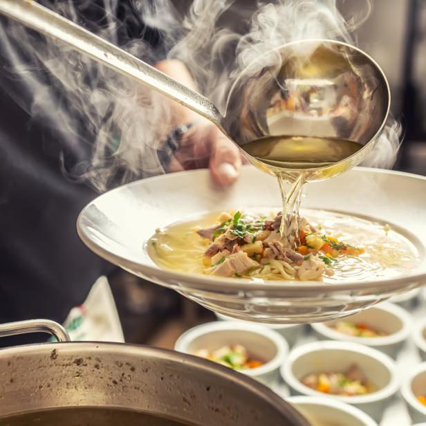 Ein Koch gießt mit einer Schöpfkelle heiße Hühnersuppe in einen Teller.