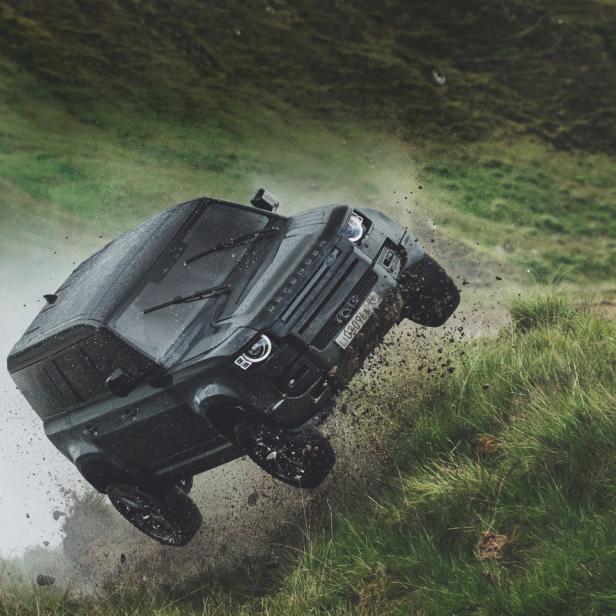 Ein schwarzer Land Rover Defender fährt über eine grüne Hügellandschaft.