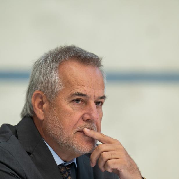 Ein Mann mit grauem Haar und Bart trägt einen dunklen Anzug und denkt nach.