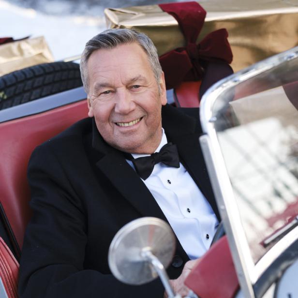 Ein lächelnder Mann im Smoking sitzt in einem Oldtimer mit Weihnachtsgeschenken im Fond.