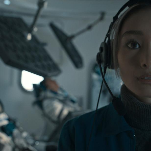Eine Astronautin mit Kopfhörern in einer Raumkapsel, während andere Astronauten im Hintergrund liegen.