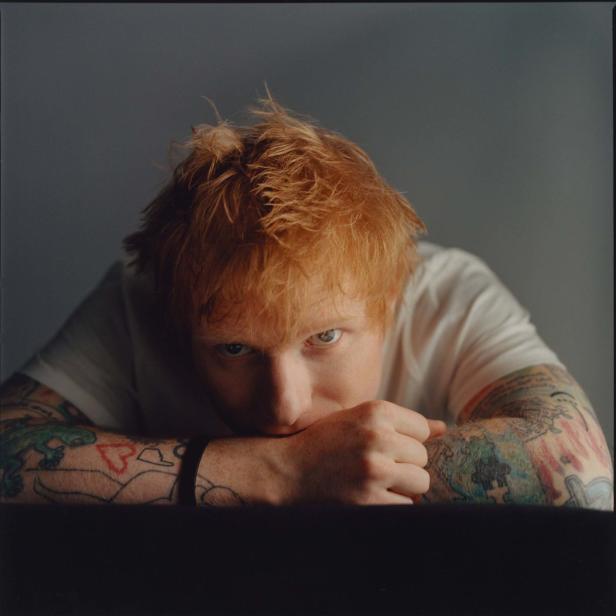 Ein Porträt von Ed Sheeran, der seine tätowierten Arme auf einem Tisch abstützt.