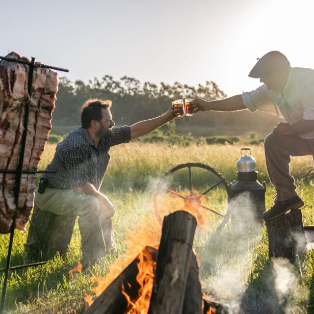 Zwei Männer stoßen mit Getränken an, während ein Asado über dem offenen Feuer brutzelt.