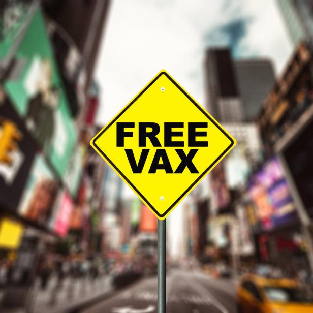 Ein gelbes Schild mit der Aufschrift „Free Vax“ vor einer belebten Stadtkulisse.