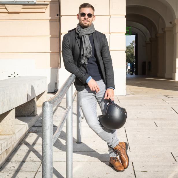 Ein Mann mit Sonnenbrille lehnt an einem Geländer und hält einen Motorradhelm.