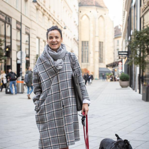 Eine Frau mit einem karierten Mantel und einem Schal geht mit ihrem Hund in einer Stadt spazieren.