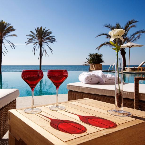Zwei rote Weingläser stehen auf einem Tisch an einem Pool mit Blick auf das Meer und Palmen.