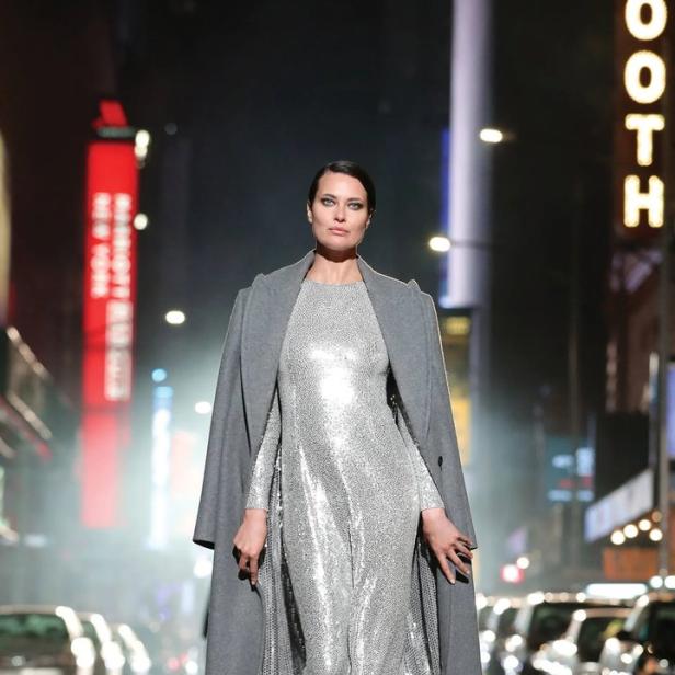 Eine Frau in einem silbernen Kleid und einem grauen Mantel geht barfuß auf einer Straße in New York.
