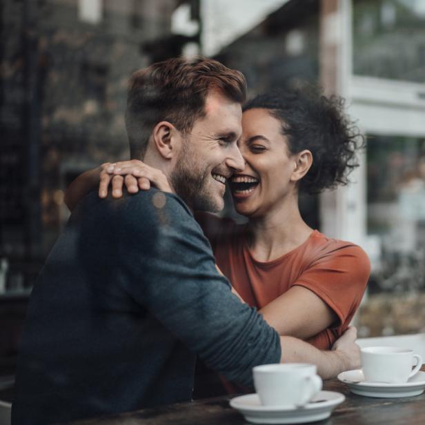 Ein lachendes Paar umarmt sich in einem Café bei zwei Tassen Kaffee.