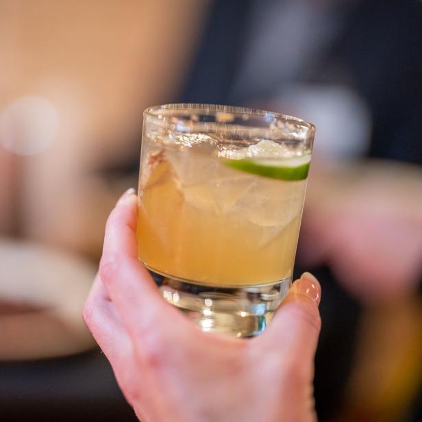 Eine Hand hält ein Glas mit einem Cocktail und einer Limettenscheibe.