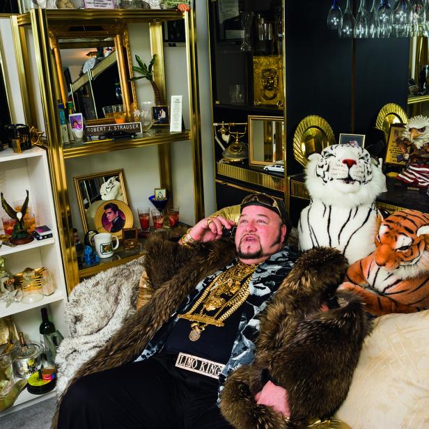 Ein Mann, vermutlich „Limo King“, sitzt mit Pelzmantel und viel Goldschmuck auf einem Sofa und telefoniert.