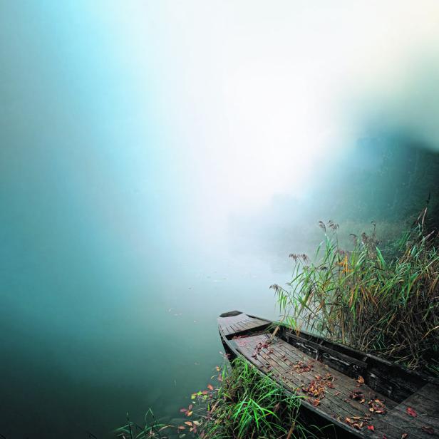 Ein verlassenes Holzboot liegt am Ufer eines nebligen Sees.